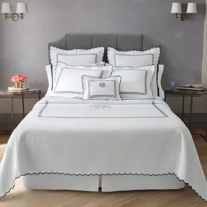 A neatly arranged white bed with scalloped, black-embroidered bedding and pillows, accented by a flower vase and book on side tables. Featuring the Classic Chain Scallop Matelassé set.