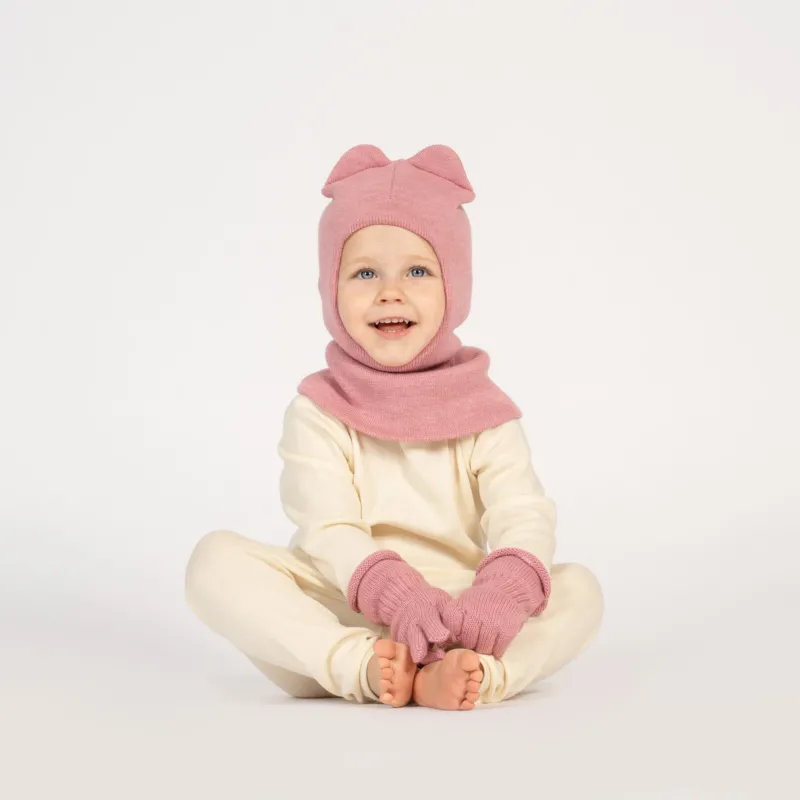 A young child with blue eyes and a wide, happy smile, is sitting cross-legged on a white surface. They are wearing a pink balaclava with ears, matching pink mittens, and a light-colored long-sleeved shirt and pants.