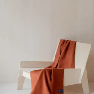 TBCo Lambswool Small Blanket in Rust Melange draped elegantly over a minimalist white chair, showcasing its soft texture and fringe