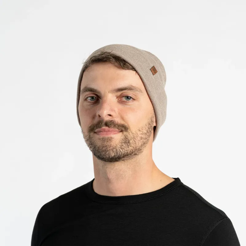 A man with light skin, blue eyes, and a beard smiles faintly while looking forward. He wears a creamy beige colored menique knit beanie with a small brown patch on the side, and a black crew-neck shirt.