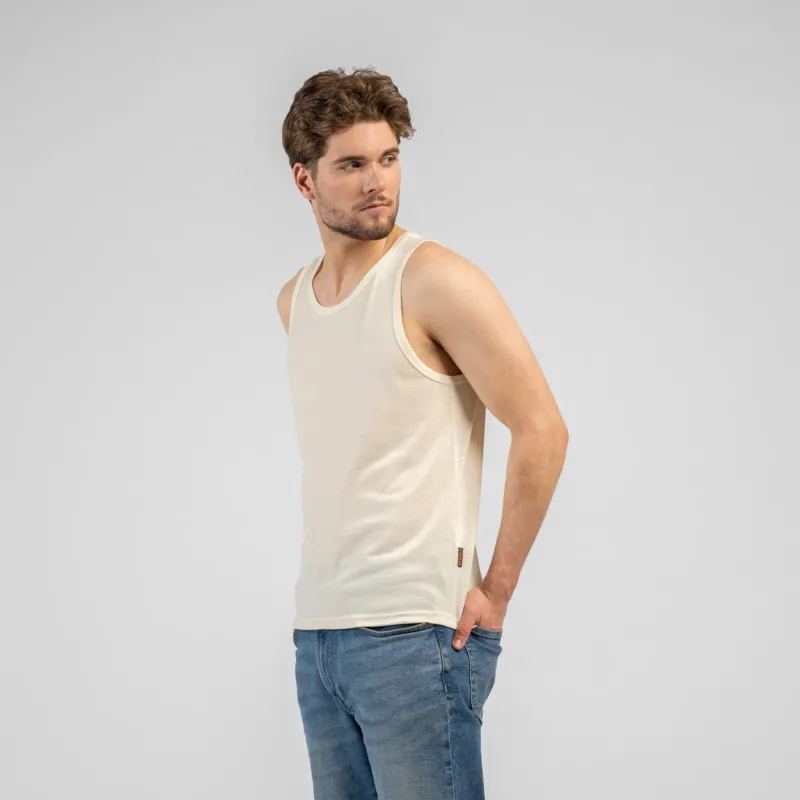 A studio portrait of a man standing and looking back over his right shoulder. He is wearing a light beige tank top and medium-wash blue jeans, with his left hand resting in his front pocket. The background is a solid light grey.