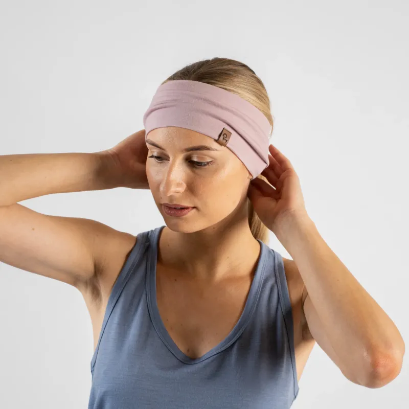 A studio portrait of a blonde woman looking downward and to the left. She is wearing a dusty blue sleeveless top and a dusty pink headband, with both hands raised behind her head to adjust the band against a light grey background.