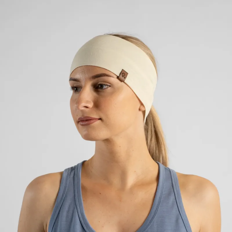A studio portrait of a blonde woman looking off to the left side of the frame. She is wearing a light cream, natural-colored headband over her pulled-back hair, featuring a small brown leather logo patch. She is wearing a dusty blue sleeveless top against a plain light grey background.